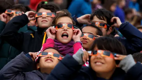 Getty Images A group of children wearing special solar viewing glasses look up at the sky during an eclipse. They are dressed in warm clothing and are gathered closely together, with expressions of awe, shock and curiosity.