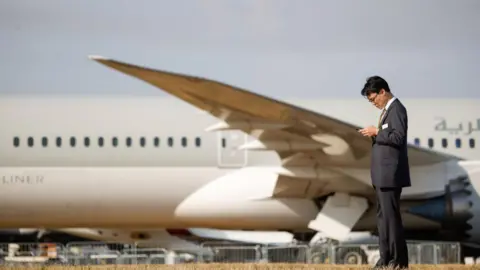 Bloomberg/Getty A man in a black suit looks at his phone while behind him is parked a Boeing 787-10 Dreamliner aircraft, operated by Qatar Airways QCSC, on the opening day of the Farnborough International Airshow in Farnborough, UK, on Monday, July 18, 2022.