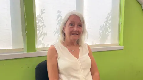 A woman in a white top smiles at the camera. The woman has white, shoulder-length hair and she is sitting in front of green wall and a window.