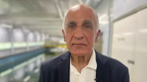 BBC News A man in a white shirt and dark jacket with some white hair in front of a long pool used for testing shipping technology.
