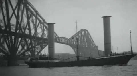 Pathe News Black and white image of the Buckau, with a black hull and two large towers sticking up from the deck.  It has just passed under the iconic Forth Bridge