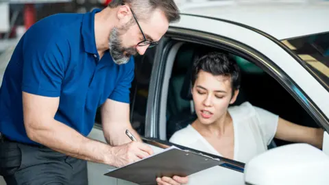 Getty Images A young woman with short black hair and a white top sits in the driver's seat of a white hatchback, and looks at paperwork on a clipboard held by the car salesman, who wears a blue t-shirt and glasses. 