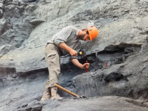 Chris Moore Chris Moore in an orange safety hat chisels at the black cliffs