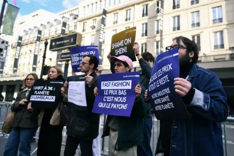 Firas Abdullah/Anadolu via Getty Images A group of protesters holding signs that say things like "fast fashion" and "shame on Shein" stand outside a row of buildings.
