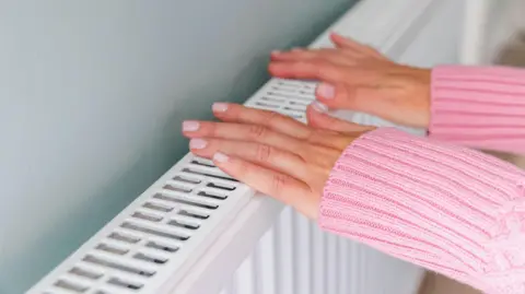 Getty Images A woman touching the top of a radiator with two hands. She is wearing a long-sleeved pink knitted jumper and has neatly manicured nails.
